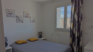 A bedroom with a bed, two nightstands, a lamp, a window with curtains, and wall art. - Sol-y-Days Grau Village, Holiday home with large terrace (Le Grau-du-Roi)
