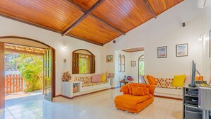 A spacious living area with a wooden ceiling, a white sofa with colorful cushions, an orange sofa, and a TV stand.