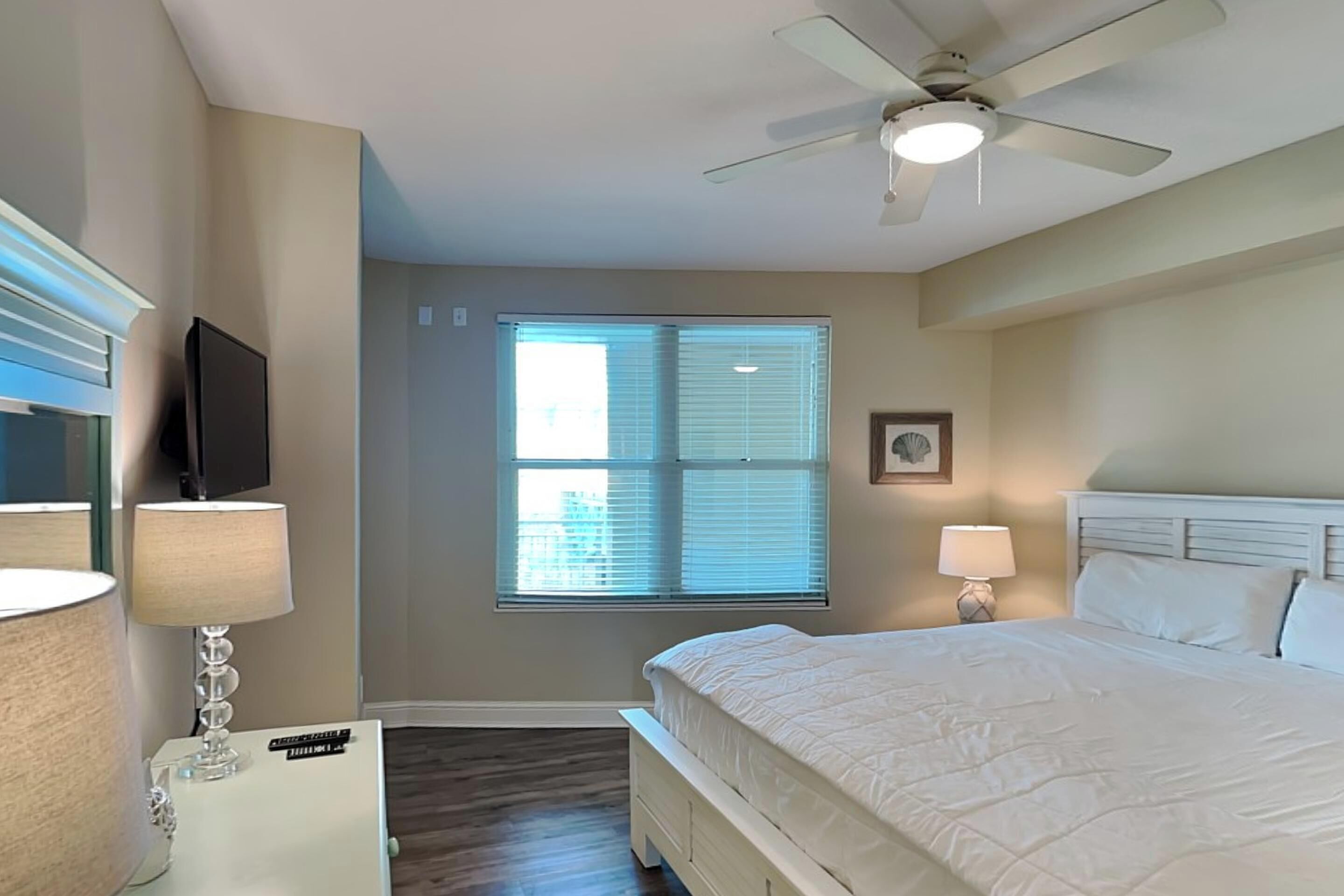 A bedroom with a bed, ceiling fan, window with blinds, and a TV mounted on the wall.