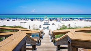 A wooden boardwalk leading to a beach with rows of blue umbrellas and lounge chairs. - Aqua #404 by Resort Collection (Panama City Beach)