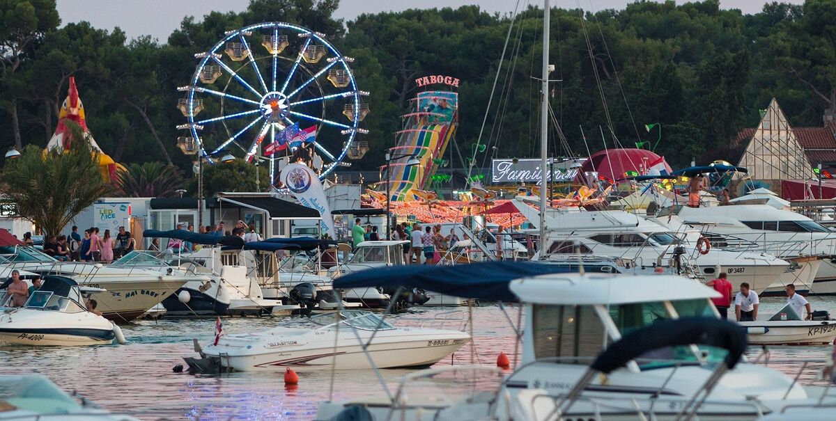 A marina with boats docked, a Ferris wheel, and a colorful amusement ride in the background.