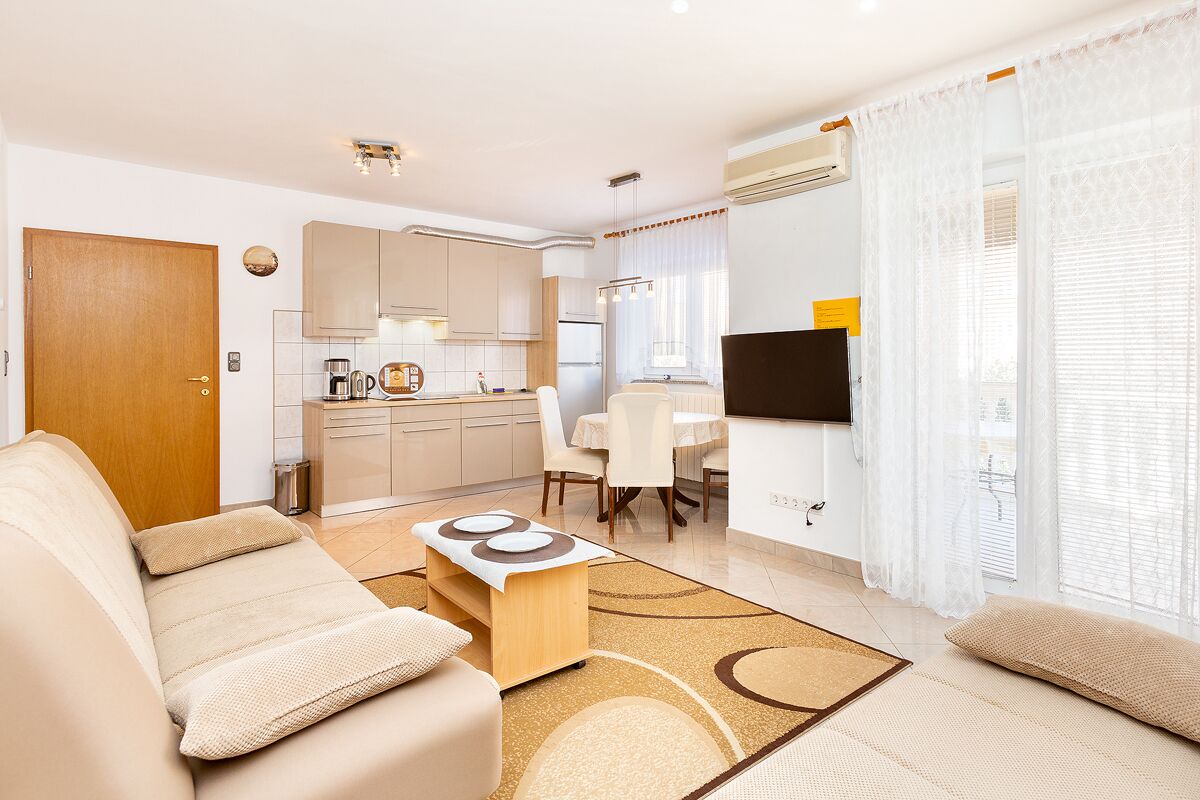 A modern living room with a sofa, coffee table, and a TV. There is a kitchen area with cabinets and a dining table.