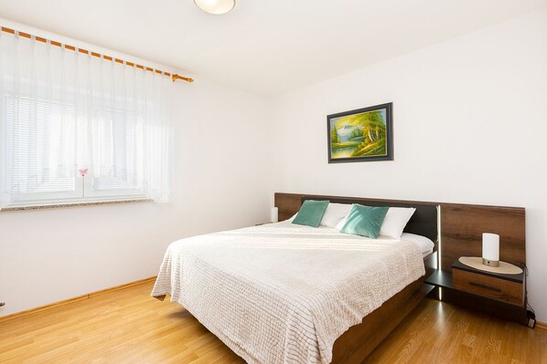 Un dormitorio con cama de madera, ropa de cama blanca, una mesita de noche con lámpara y un cuadro en la pared.