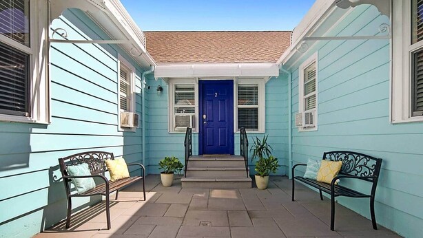 Un bâtiment de couleur bleu clair, avec une porte bleue, deux bancs et des plantes en pot.