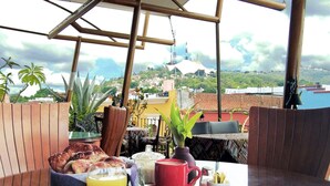A breakfast setup with bread, fruit, and beverages on a table under a pergola with a city view. - Boutique Hotel Casa Cid de León (Oaxaca)