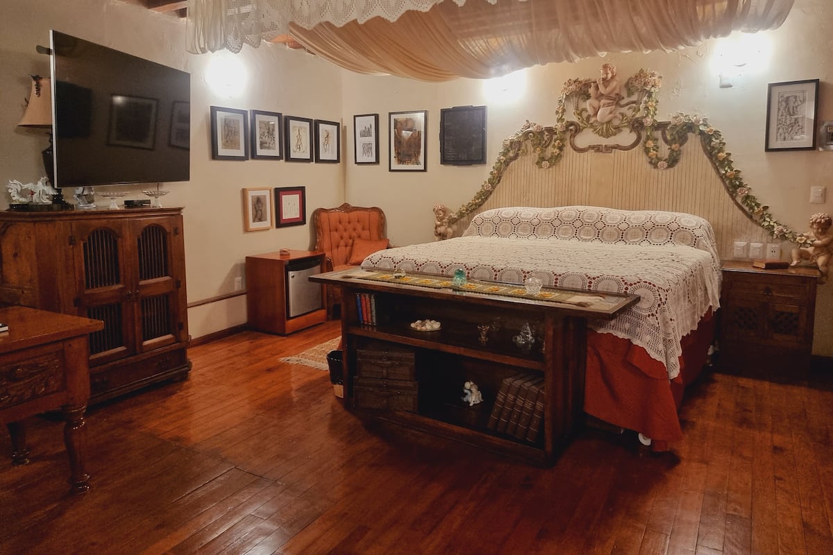 A cozy bedroom with a wooden floor, a large bed with a decorative headboard, a wooden nightstand, a dresser, and a television on the left wall.