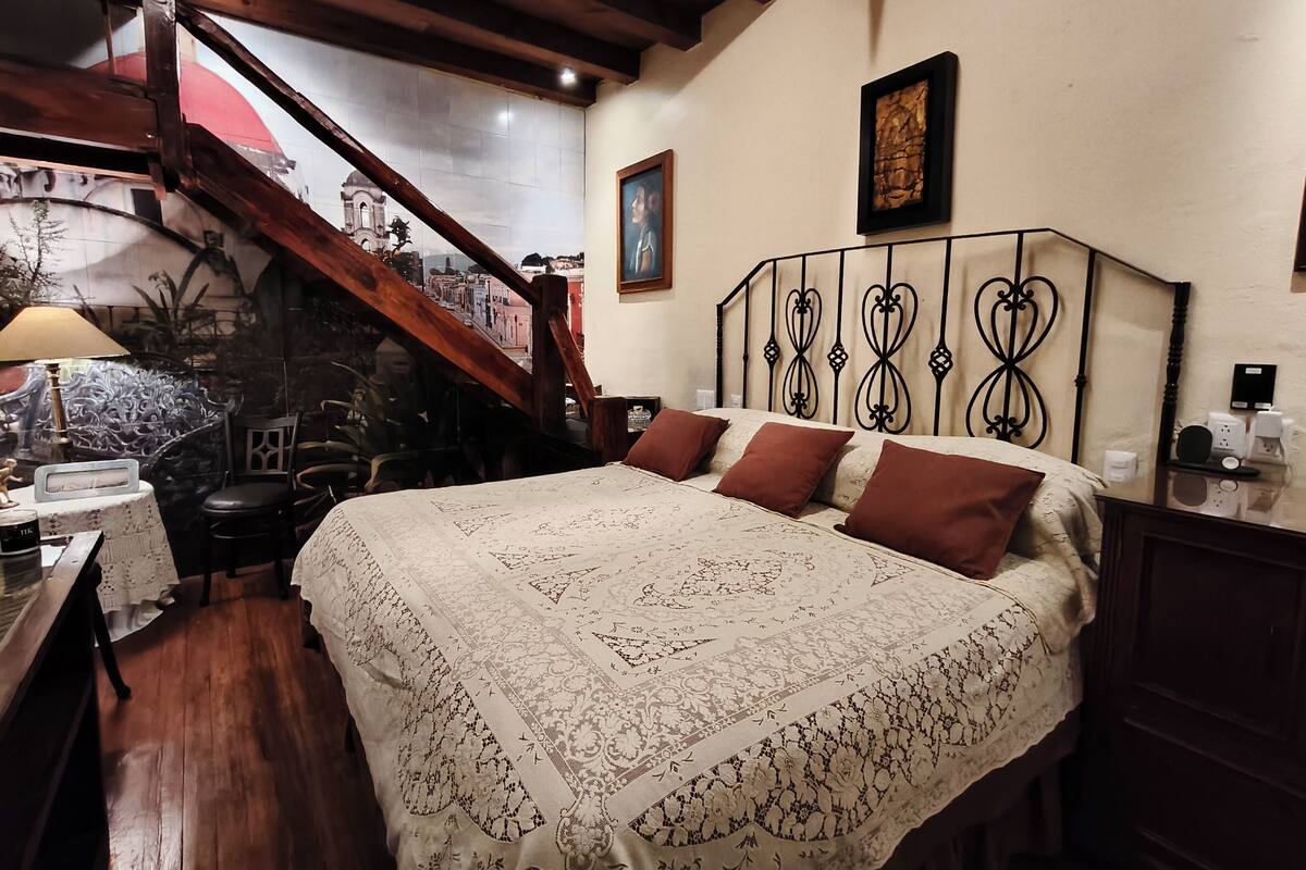 A bedroom with a large bed, wooden beams, a staircase, and a cityscape mural on the wall.
