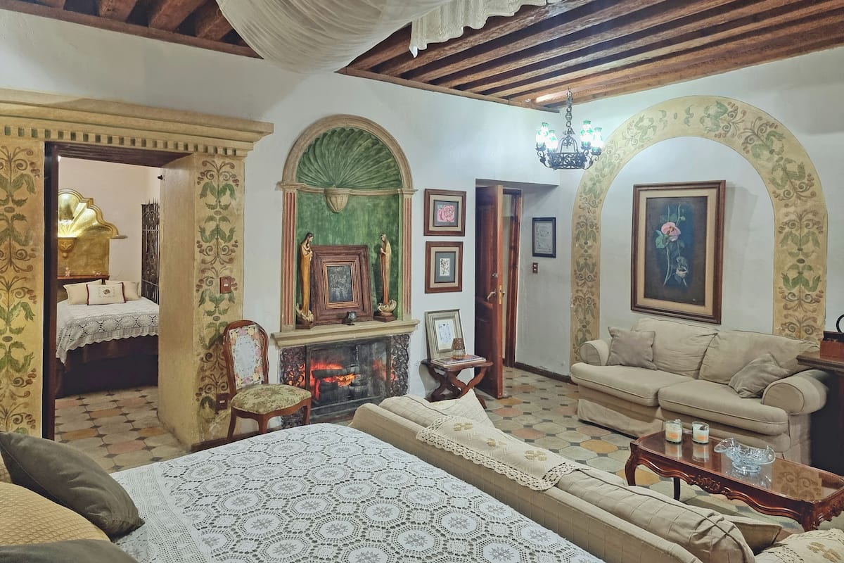 A cozy living room with a fireplace, a sofa, a dining table, and a bed with a patterned bedspread.