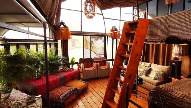 A cozy room with a wooden floor, a red sofa, a beige sofa, and a ladder. There are plants and a window with a view of buildings. - Boutique Hotel Casa Cid de León (Oaxaca)