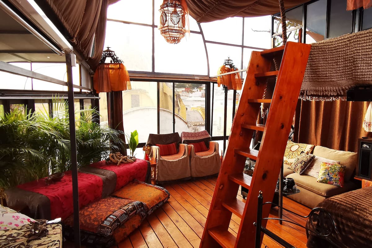 A cozy room with a wooden floor, a red sofa, a beige sofa, and a ladder. There are plants and a window with a view of buildings.