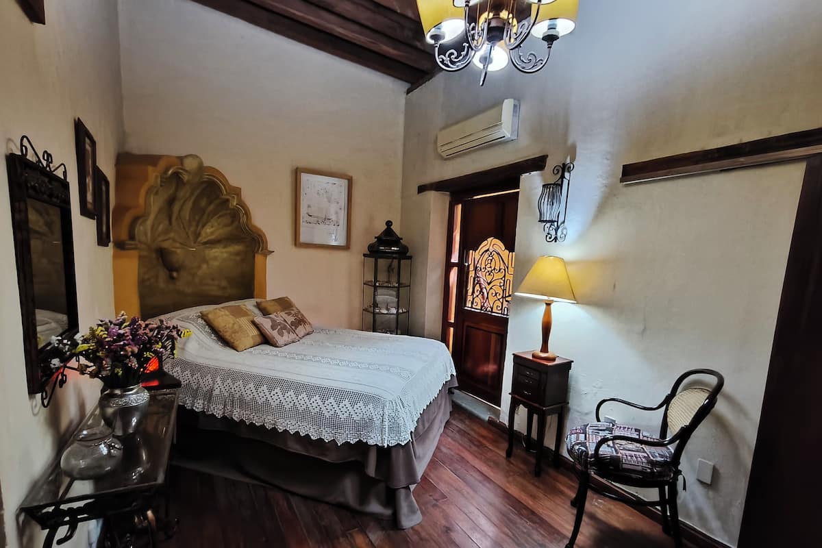 A bedroom with a bed, a nightstand, a chair, a lamp, a mirror, and a wall-mounted air conditioner.