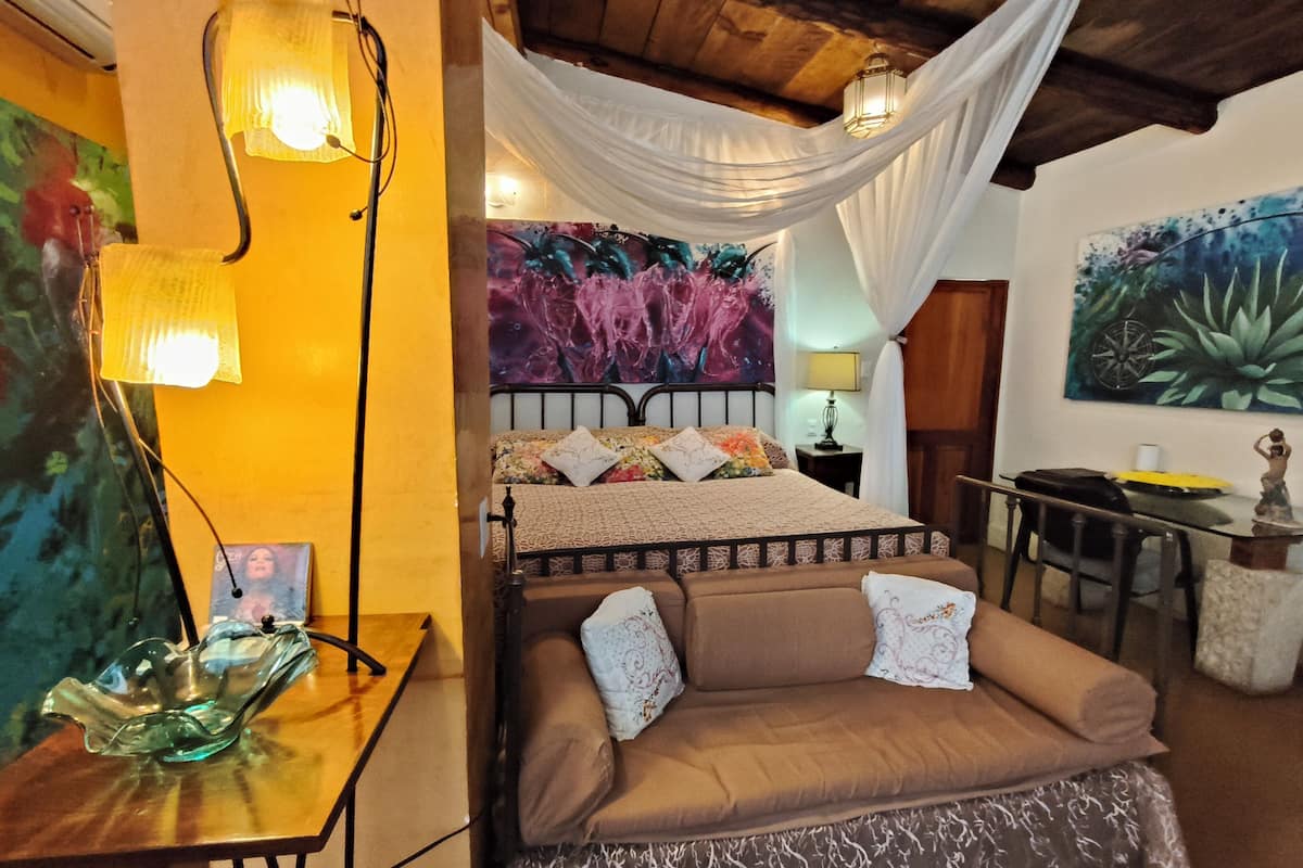 A bedroom with a bed, a bedside table, a lamp, a chair, a desk, and a painting on the wall.