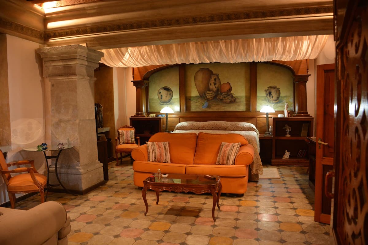 A hotel room with a large bed, an orange sofa, a wooden coffee table, and a stone pillar.