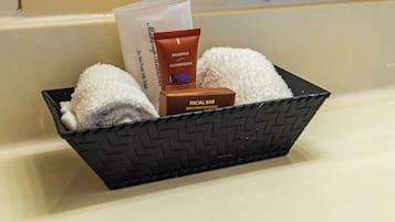 A black woven basket containing a facial bar, a shampoo tube, and two white towels on a beige countertop.