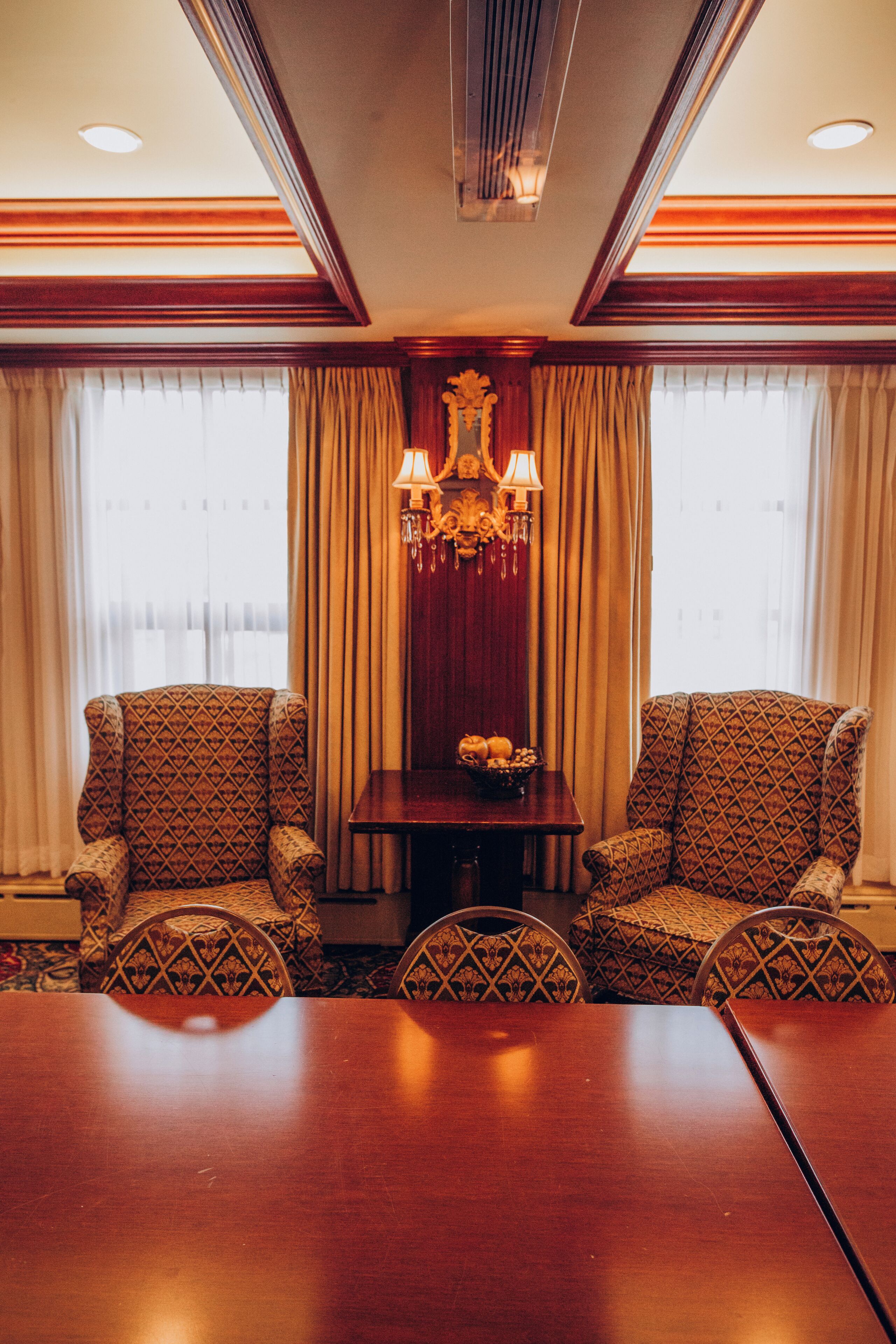 Un salón de conferencias con una mesa de madera, sillones estampados, una araña y cortinas.