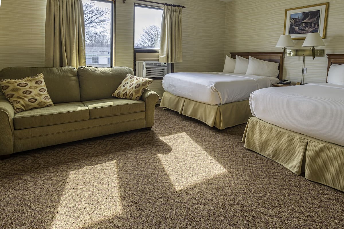 A hotel room with a sofa, two beds, a window with curtains, a radiator, and a lamp.