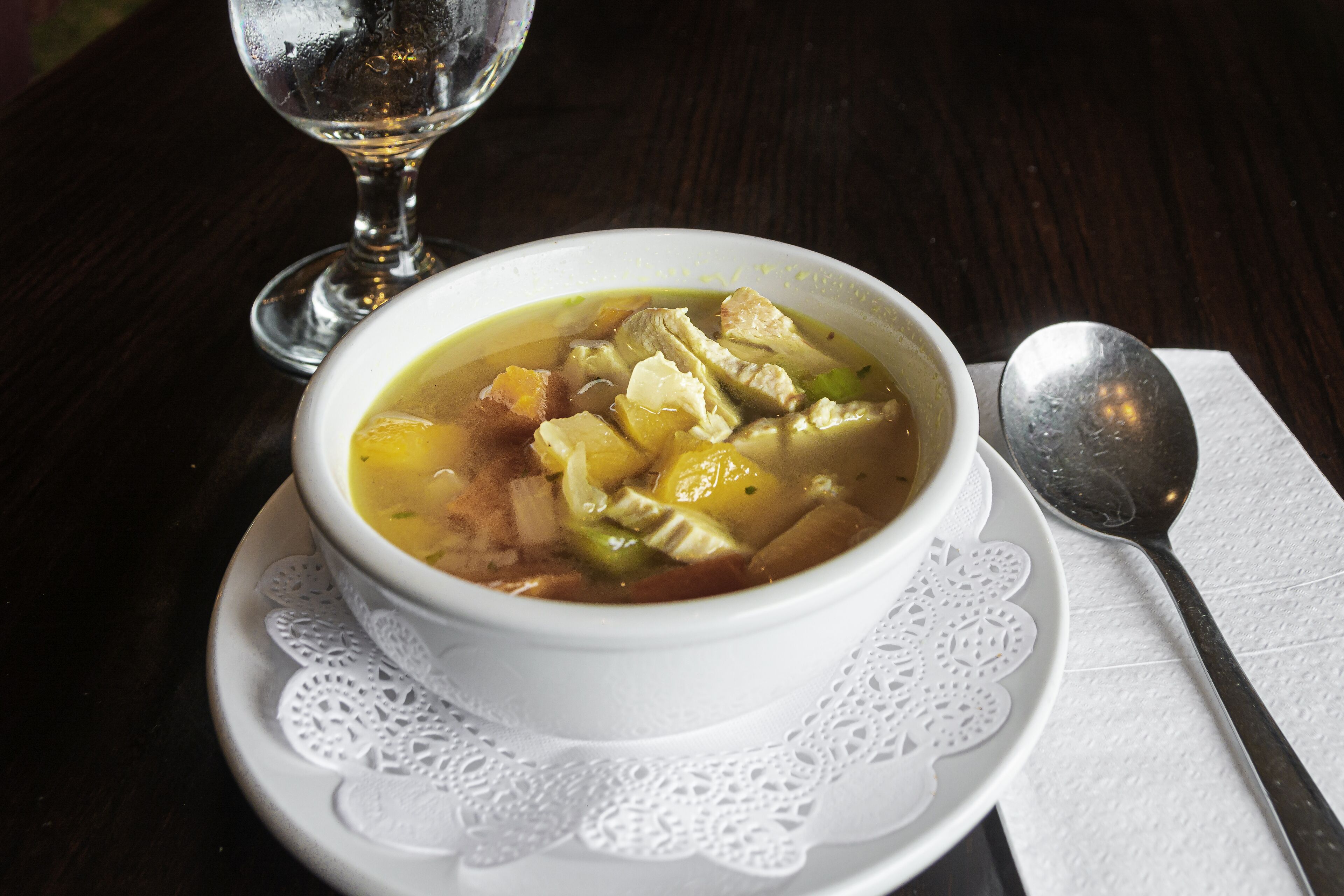 Un tazón de sopa sobre un plato blanco, junto a una cuchara y un vaso de agua, todo sobre una mesa de madera oscura.