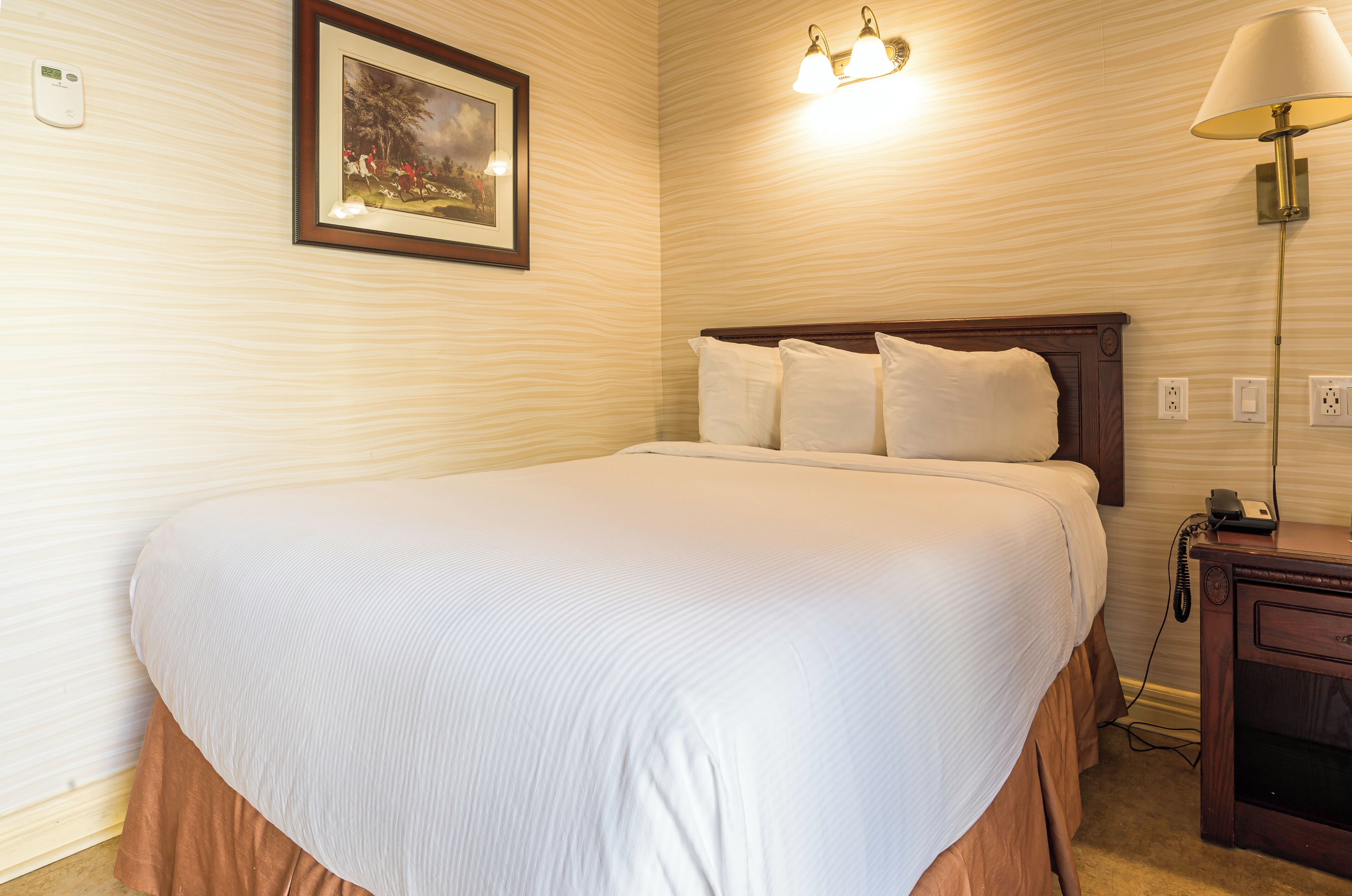Una cama bien tendida con cabecera de madera, una lámpara de noche y un cuadro en la pared.