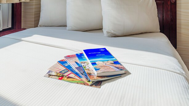 A neatly made bed with white linens and pillows, a stack of travel brochures, and a wooden headboard. - The Guv’nor (St. John's)