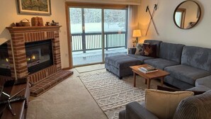 A cozy living room with a fireplace, a gray sofa, a patterned rug, and a view of a snowy deck through a glass door.