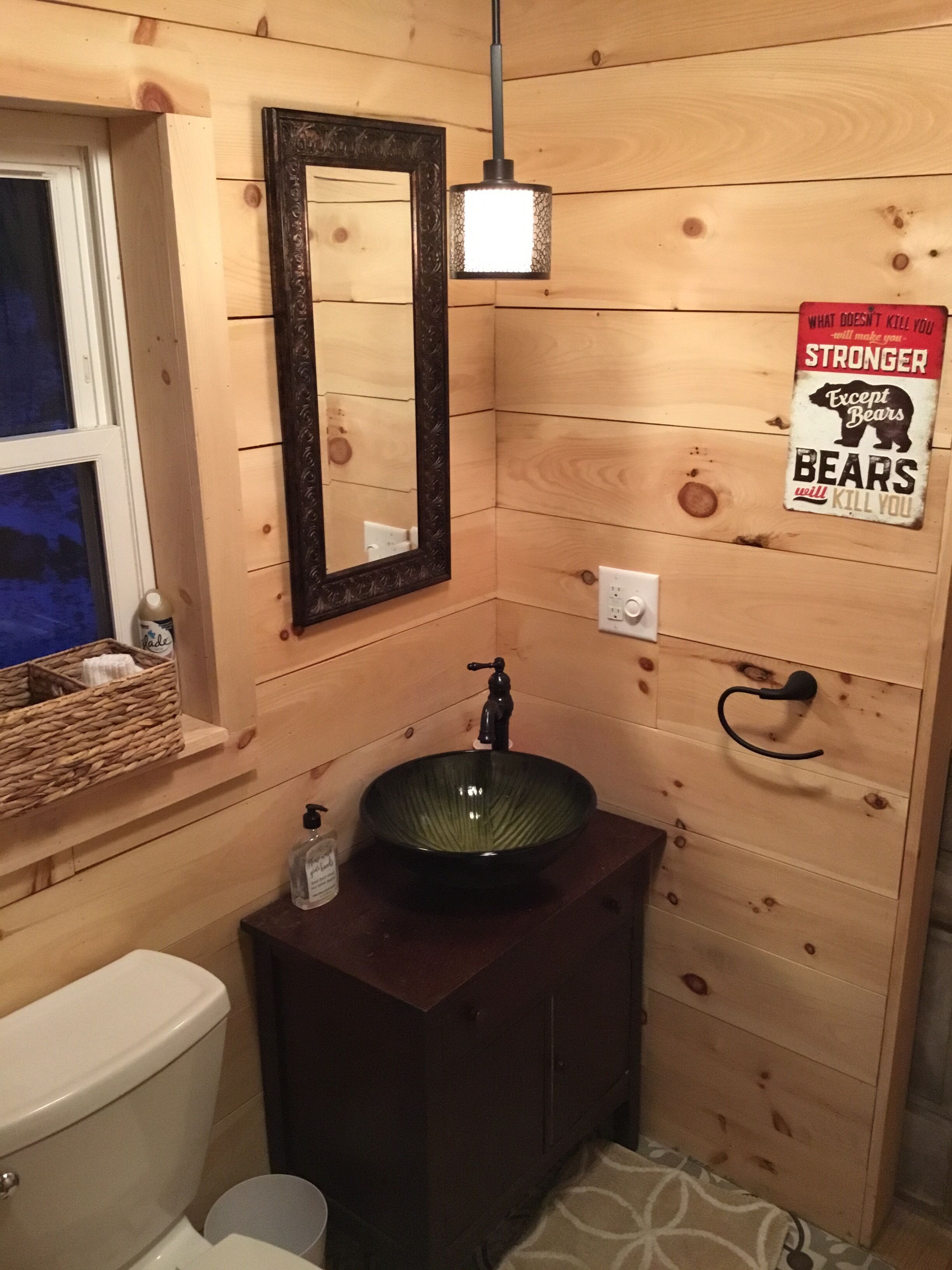 Baño de cabaña de madera con lavamanos verde, un espejo, una lámpara colgante, una ventana y un cartel con motivos de oso.