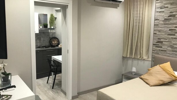 Ein kleines Hotelzimmer mit Bett, einem kleinen Nachttisch, einer Wand-Klimaanlage und Blick auf den Küchenbereich.