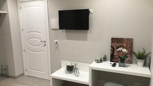 Ein Zimmer mit einer weißen Tür, einem an der Wand befestigten Flachbildfernseher, einem weißen Schreibtisch mit einer Blumenkorb sowie einem kleinen Hocker.
