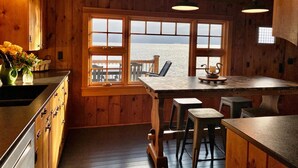 A wooden cabin interior with a kitchen area, large windows overlooking the sea, and a bar counter.