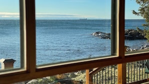A view of the ocean through a window with a wooden frame, a rocky shoreline, and a distant ship.