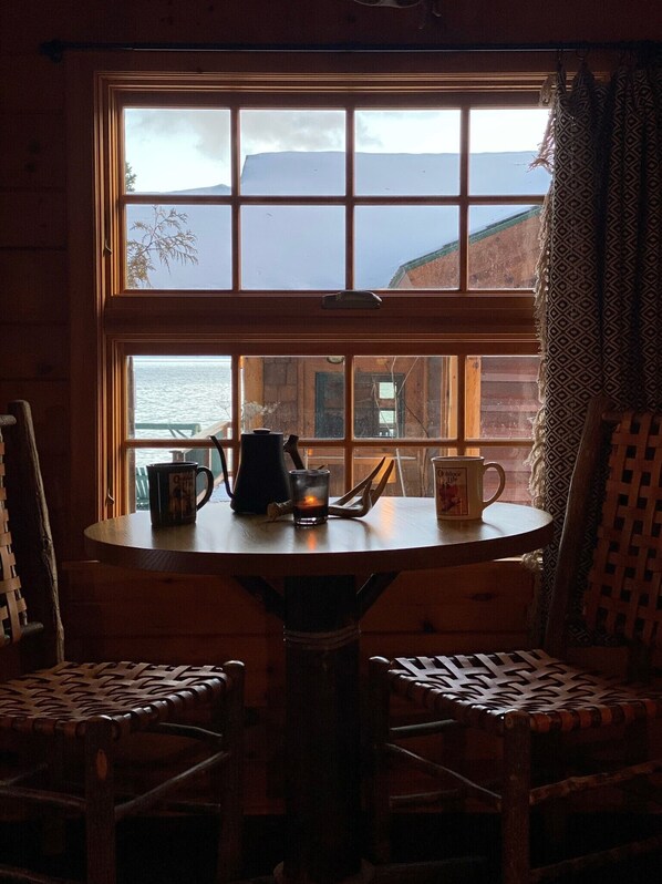 A wooden cabin with a large window overlooking a body of water and mountains. Inside, there is a round wooden table with two chairs, a coffee pot, and a mug.