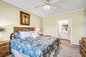 A bedroom with a bed, nightstands, a ceiling fan, a wooden dresser, a mirror, and a door leading to a bathroom. - Legends Golf Resort Condos by Escape (Myrtle Beach)