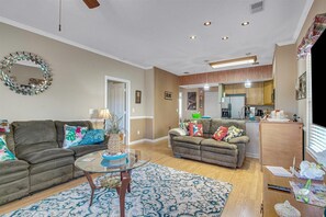 A living room with a sectional sofa, a glass coffee table, and a patterned rug. There is a kitchen area with wooden cabinets and a refrigerator. - Legends Golf Resort Condos by Escape (Myrtle Beach)