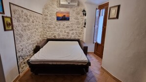 A narrow room with a bed, stone walls, a small window, and a door.
