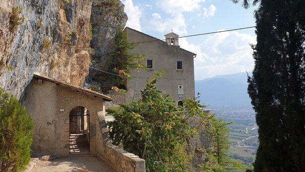 A stone building with a bell tower, a narrow passageway, and a view of a valley.