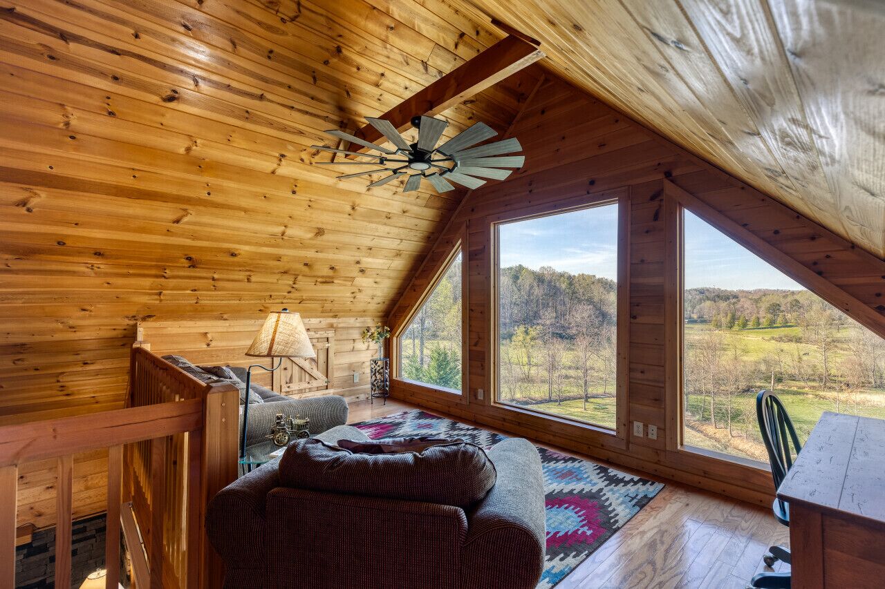 A wooden cabin interior with a sloped ceiling, a ceiling fan, a large window offering a view of a forested landscape, a cozy seating area with a patterned rug, and a small table.