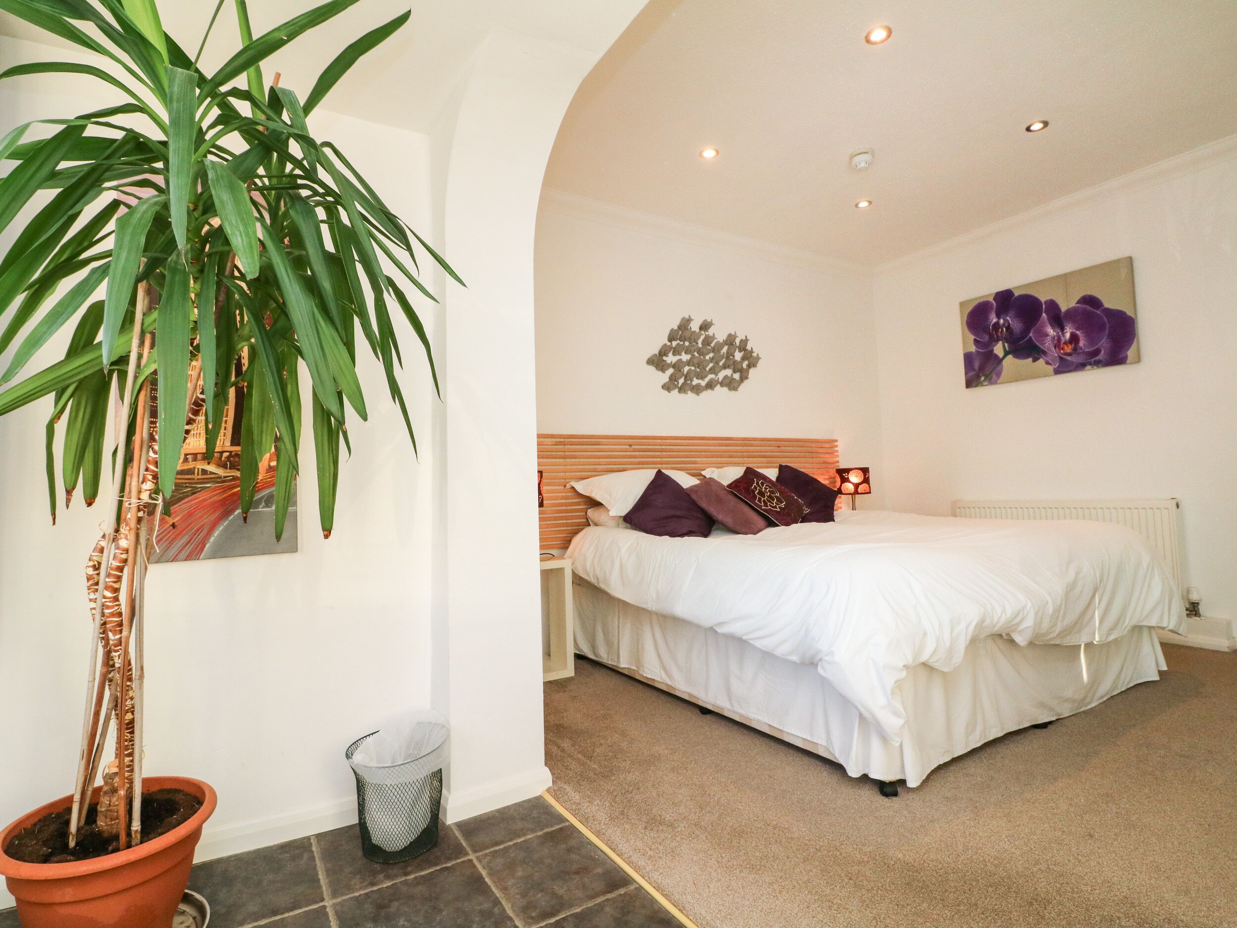 Un dormitorio con una cama, una planta en maceta, un cubo de basura y una pared decorada con una pintura de flores.