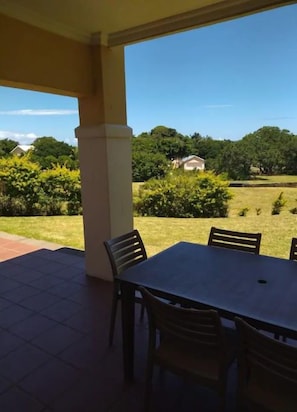 Une terrasse couverte, avec une table et des chaises, donnant sur une pelouse arborée et une maison située au loin.