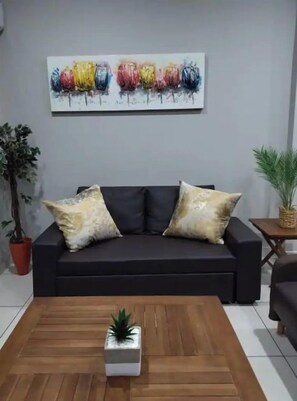 Un salon moderne avec un canapé foncé, deux coussins décoratifs, une table basse en bois agrémentée d’une petite plante, et un tableau abstrait aux couleurs vives accroché au mur.