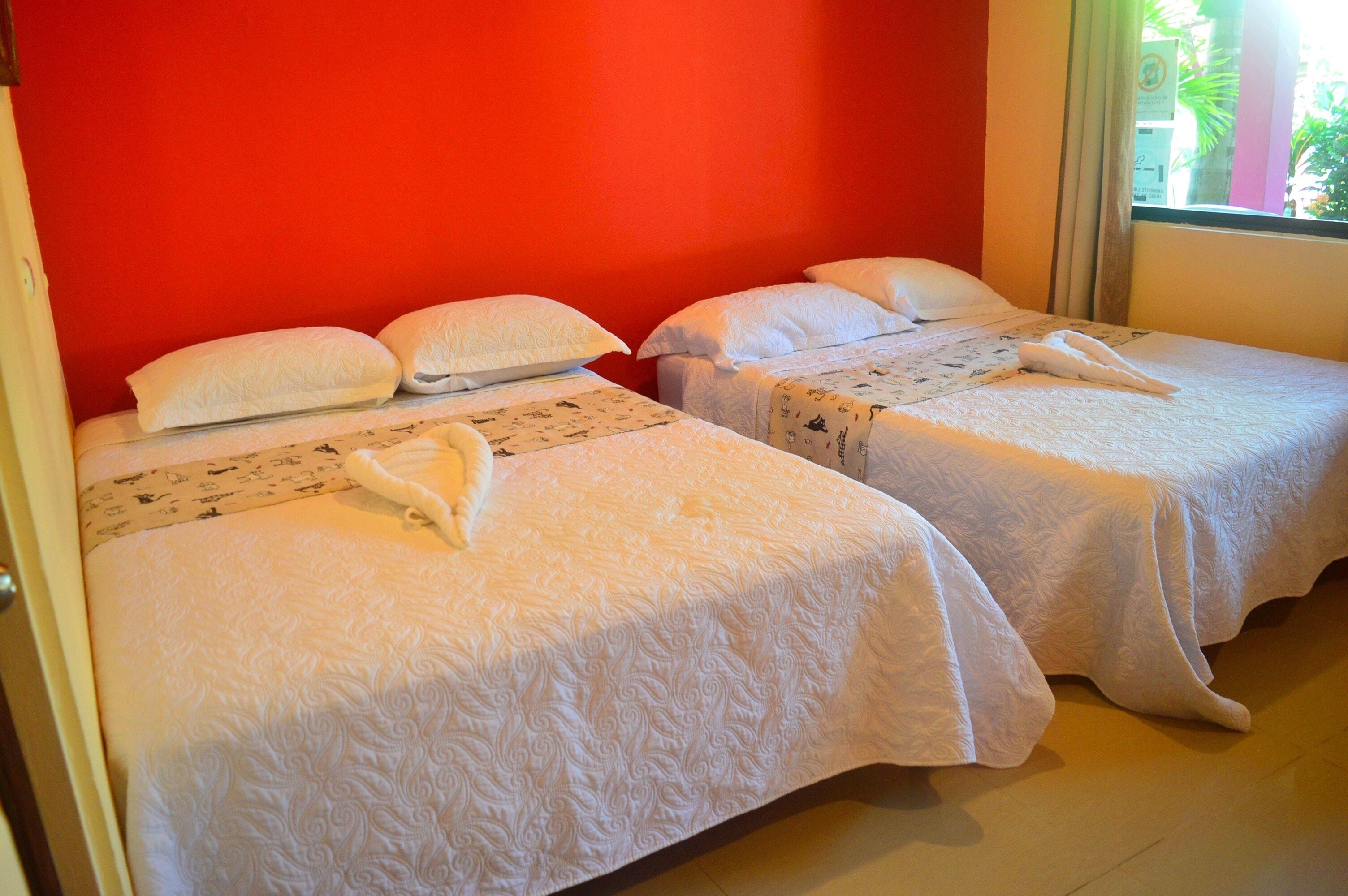 Dos camas individuales con sábanas y almohadas blancas, una pared roja y una ventana con vista a follaje.