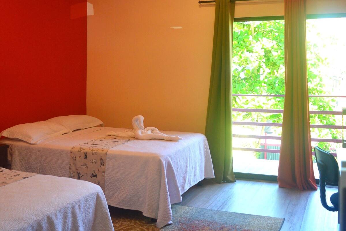 A hotel room with two beds, a desk, and a view of greenery outside.