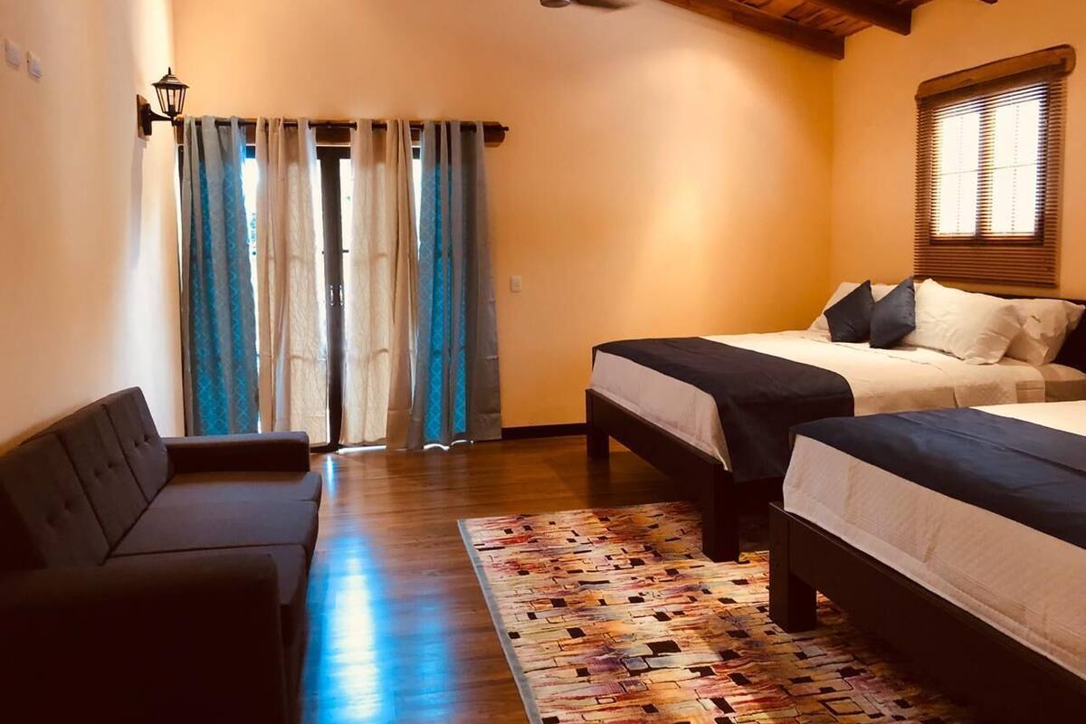A bedroom with two beds, a sofa, a ceiling fan, and a window with blinds.