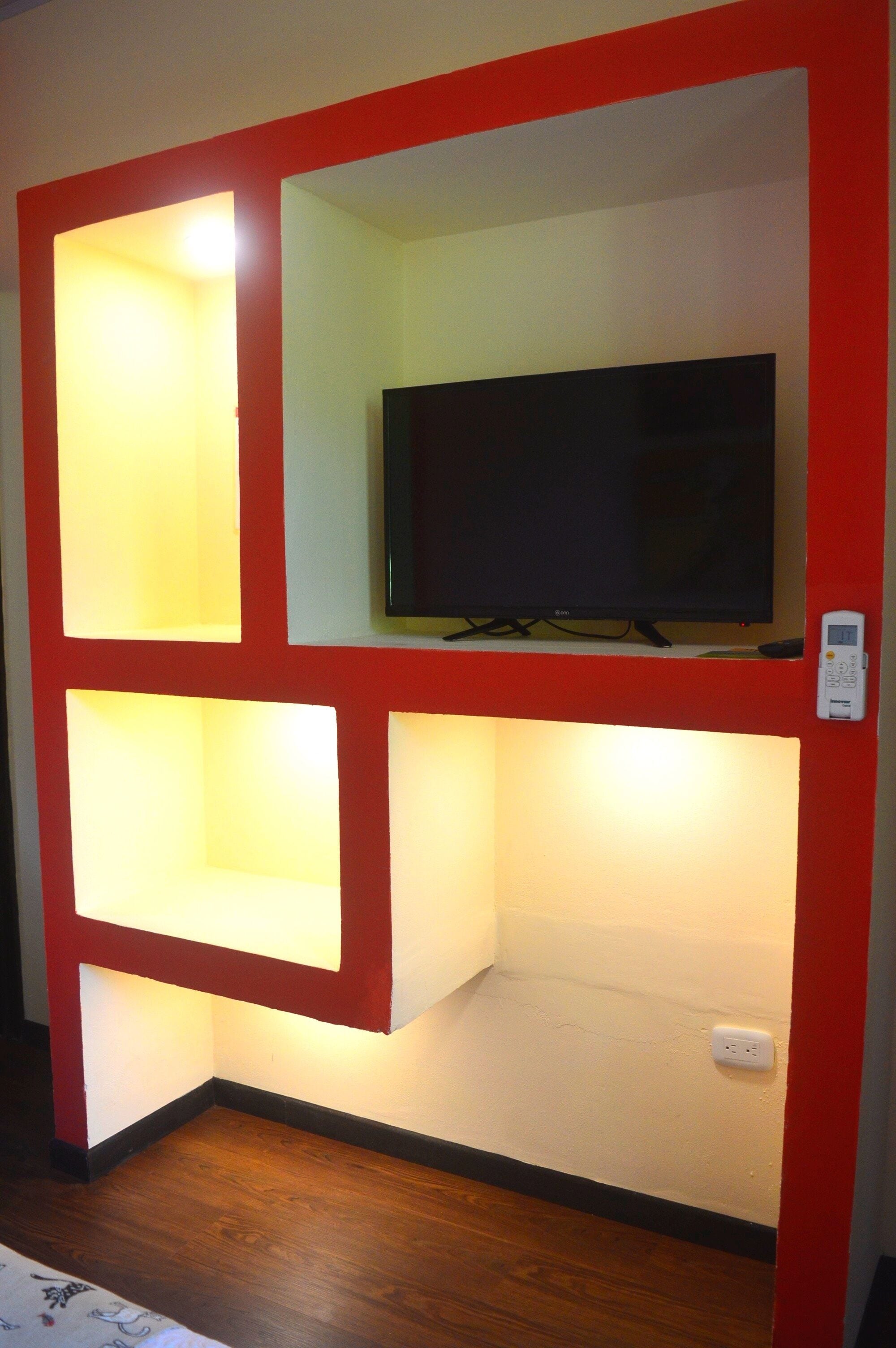 Een kamer met een rode lijst aan de wand, een televisie op een plank, een witte muur en een houten vloer.