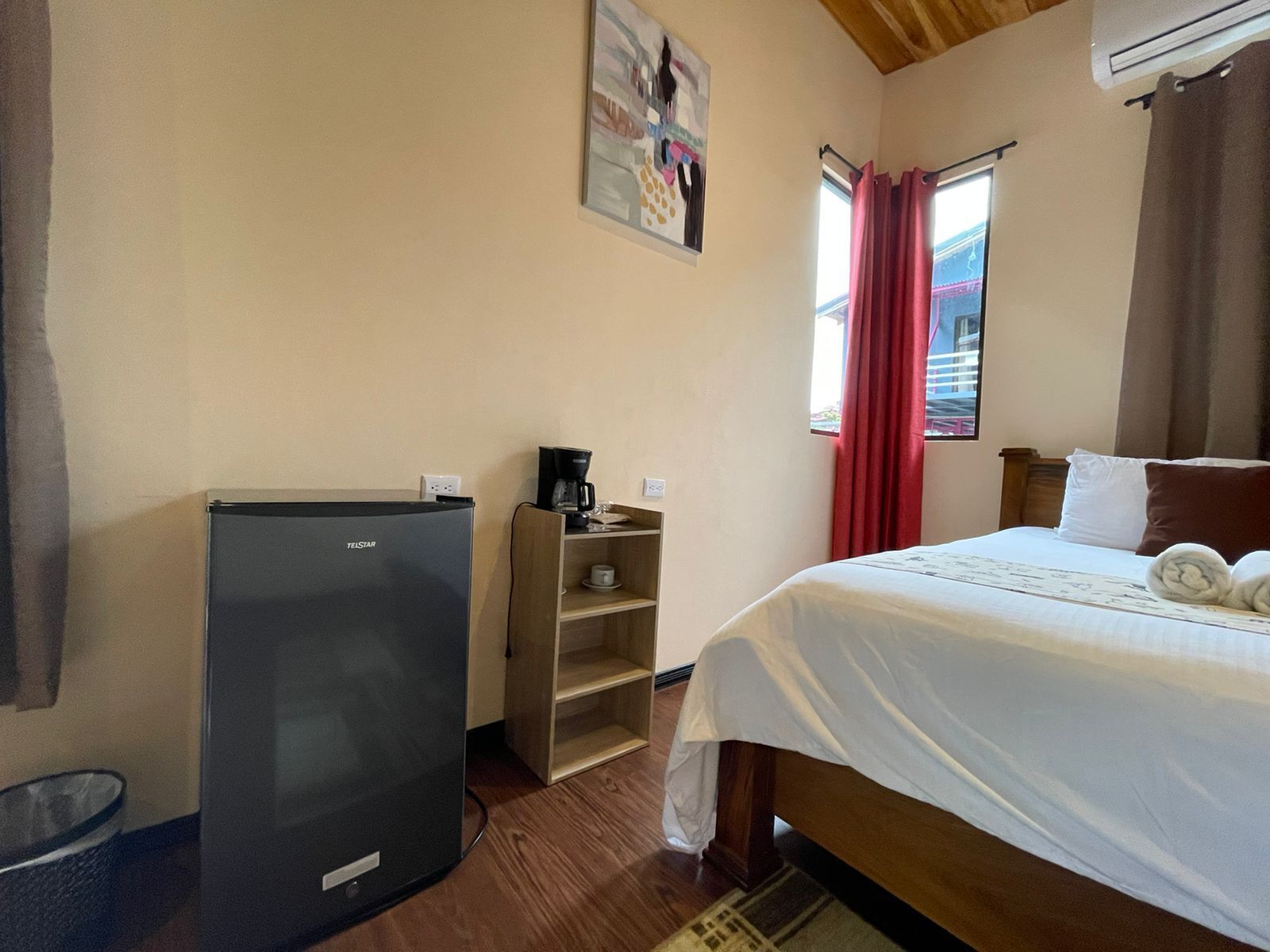 A hotel room with a bed, a refrigerator, a coffee maker, and a window with red curtains.