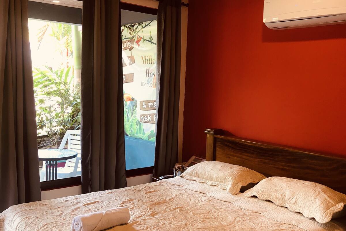 A hotel room with a large bed, a wooden headboard, a window with curtains, and an air conditioning unit on the wall.