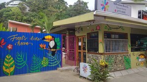 A colorful building with a mural of a toucan and tropical plants, a sign for Millennium Hostels, and a small shop with a sign for a local business. - White Sands Hotel (Quepos)