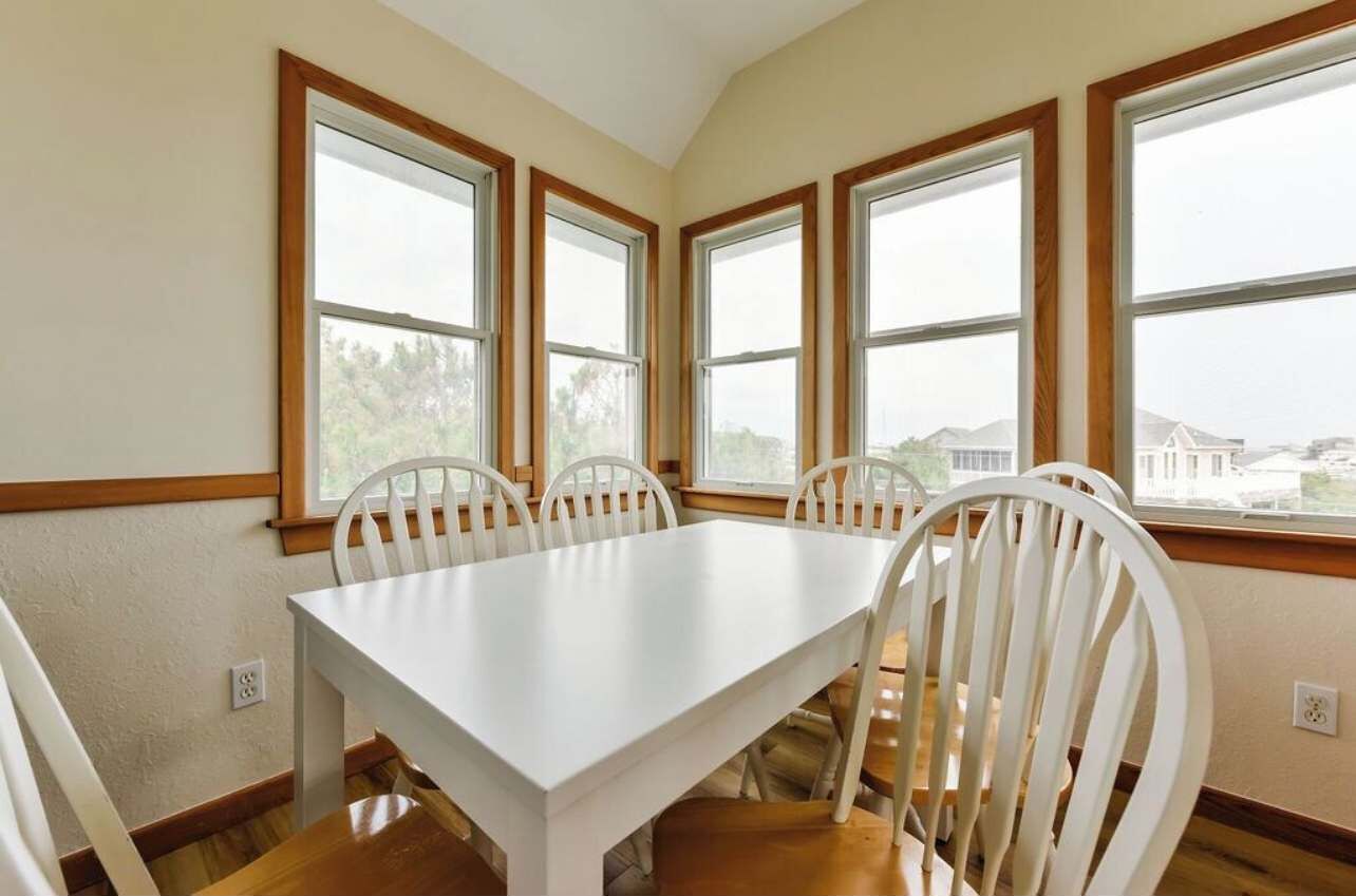 A dining room with a white table and chairs, large windows with wooden frames, and a view of houses and trees.