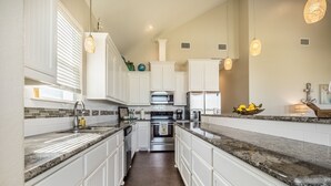 Una cocina moderna con armarios blancos, encimeras de granito, electrodomésticos de acero inoxidable e iluminación colgante.