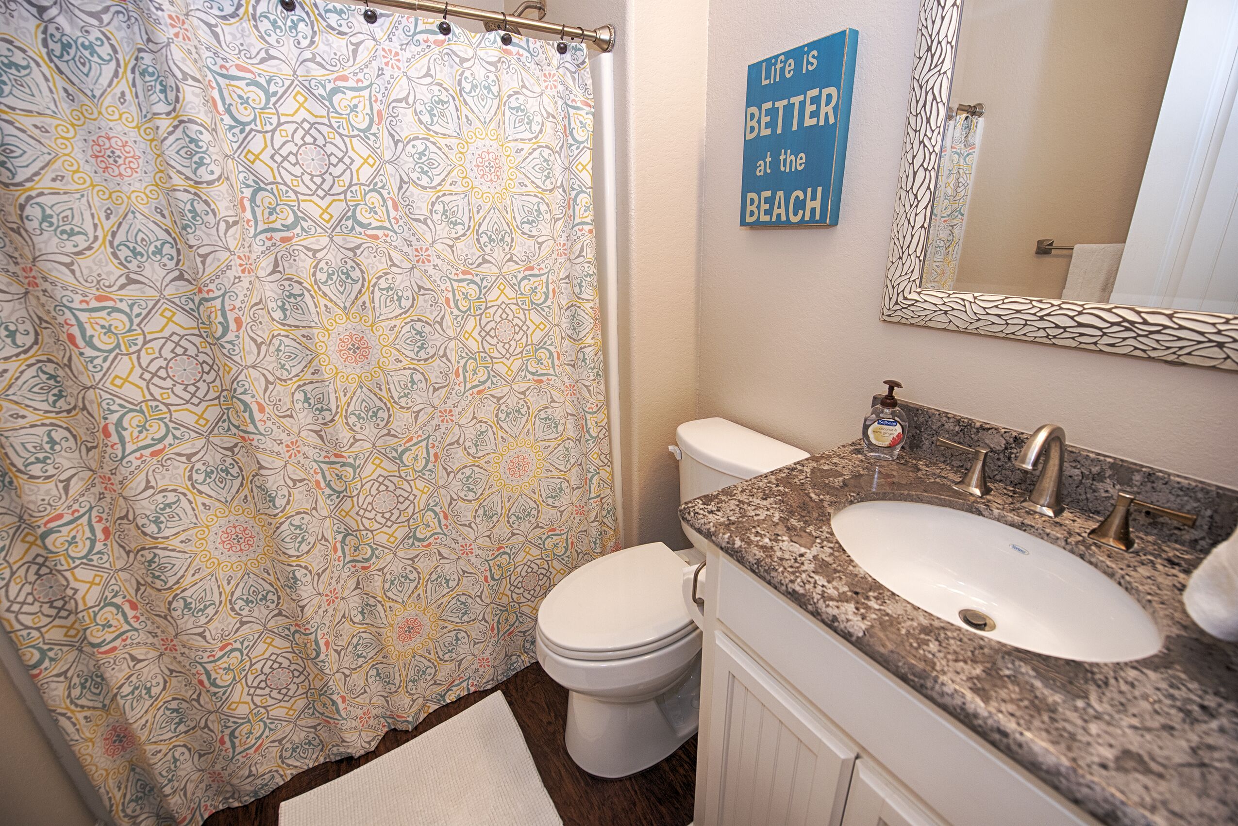 A bathroom with a patterned shower curtain, a toilet, a sink with a granite countertop, and a mirror.