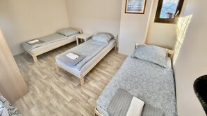 Ein kleiner, sauberer Raum mit drei Einzelbetten, einem kleinen Tisch und einem gerahmten Bild an der Wand.