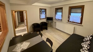 Ein gemütlicher Wohnraum mit Esstisch, zwei Stühlen, einer Couch, einem Fernseher und einem Fenster mit Vorhängen.